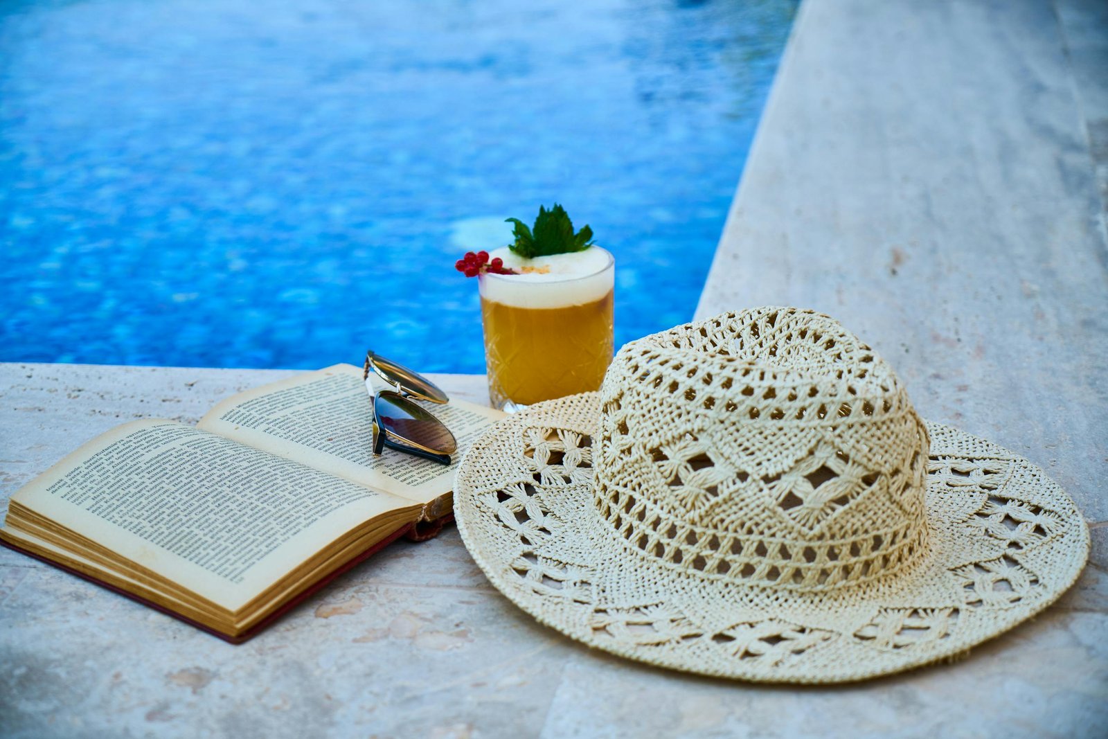 Beige Straw Hat, Book, Sunglasses, and Drink Beside Pool